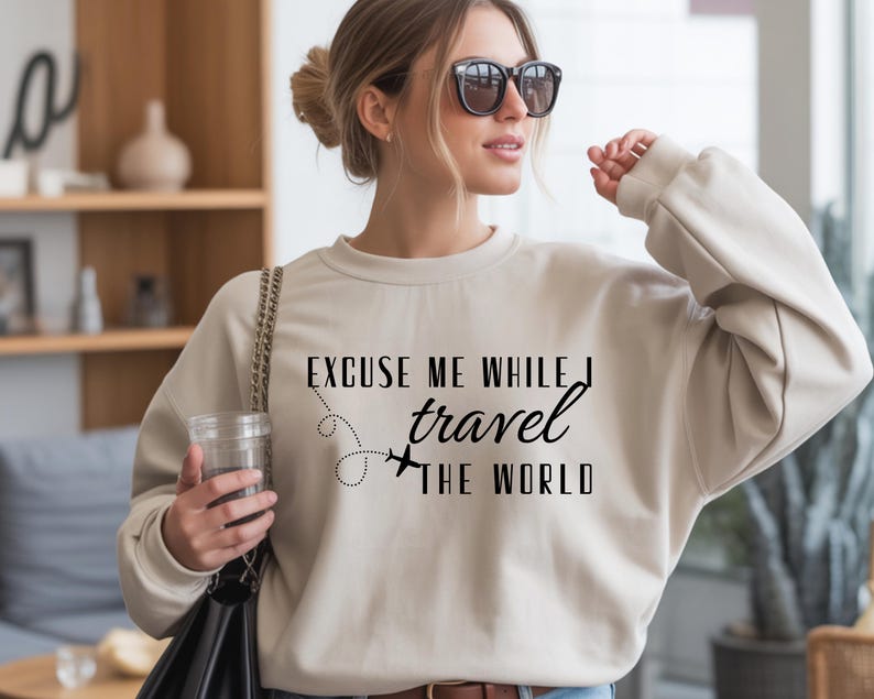 May include: A cream-colored sweatshirt with the text "EXCUSE ME WHILE I travel THE WORLD" in black. The design includes a dotted line and airplane graphic. The sweatshirt is a casual travel-themed apparel item.