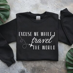 May include: Black crewneck sweatshirt with the text "Excuse Me While I Travel The World" in white. The design includes a dotted line and airplane graphic. The sweatshirt is a casual travel-themed apparel item.