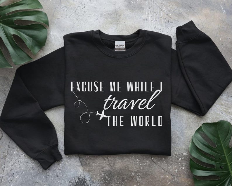 May include: Black crewneck sweatshirt with the text "Excuse Me While I Travel The World" in white. The design includes a dotted line and airplane graphic. The sweatshirt is a casual travel-themed apparel item.
