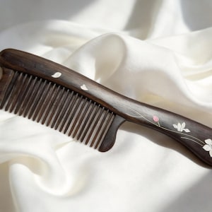 May include: A dark brown wooden comb with fine teeth and a curved handle. The handle is decorated with white and pink floral designs. The comb is resting on a white, silky fabric.