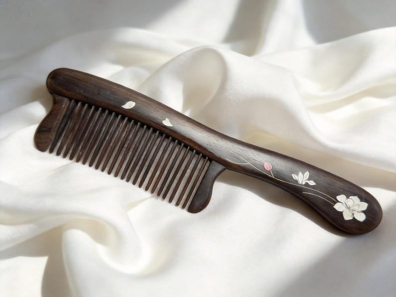 May include: A dark brown wooden comb with fine teeth and a curved handle. The handle is decorated with white and pink floral designs. The comb is resting on a white, silky fabric.