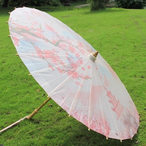 Puede incluir: Un parasol de papel blanco con mango de bambú, abierto sobre hierba verde. El parasol presenta un delicado diseño floral en rosa y rojo, con una ilustración de pagoda.