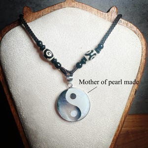 Könnte beinhalten: Eine schwarze Kordelkette mit einem Yin-Yang-Anhänger aus Perlmutt. Der Anhänger ist rund und hat ein schwarz-weißes Design. Die Kette hat außerdem schwarze Perlen und dekorative weiße und schwarze Perlen.
