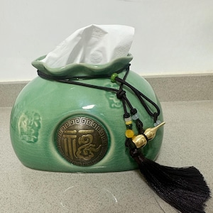 May include: A jade green ceramic tissue box with a round shape and a decorative tassel. The box features a bronze emblem with Chinese characters and a black cord with beads. White tissues are visible.