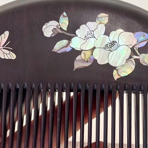 May include: A dark brown wooden comb with a decorative design. The comb features a butterfly and floral design inlaid with iridescent mother-of-pearl. The comb's teeth are evenly spaced and finely crafted.
