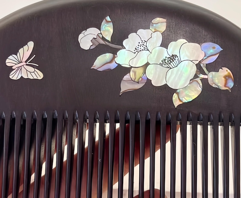 May include: A dark brown wooden comb with a decorative design. The comb features a butterfly and floral design inlaid with iridescent mother-of-pearl. The comb's teeth are evenly spaced and finely crafted.