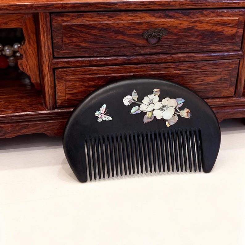 May include: A black wooden comb with a curved handle and fine teeth. The comb is decorated with a butterfly and floral design inlaid with mother-of-pearl. The comb is displayed on a white surface with a wooden cabinet in the background.