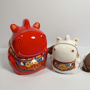 May include: Two ceramic horse figurines. One is bright red, the other is white. Both have gold accents on their ears and a decorative collar with a gold coin. The red horse has a glossy finish, while the white horse has a matte finish.