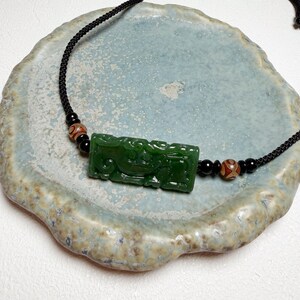 May include: A dark green jade pendant necklace with a rectangular carved jade pendant. The pendant is suspended on a black cord with black and patterned beads. The necklace is displayed on a light blue and beige ceramic plate.