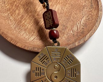 Puede incluir: Un colgante octogonal de color latón con caracteres chinos y un símbolo Yin-Yang. Cuelga de un cordón negro con una cuenta de madera roja y una pieza rectangular con texto dorado. El colgante está sobre un plato de madera.