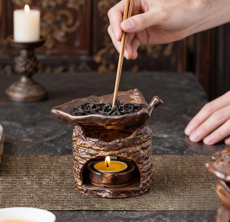 May include: A brown ceramic tea warmer with a leaf-shaped top filled with tea leaves. A lit candle sits inside the warmer, and a person is using chopsticks to handle the tea leaves. The warmer has a textured, bark-like design.