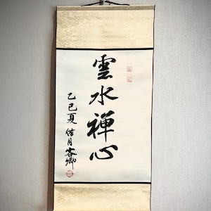Könnte beinhalten: Eine traditionelle japanische Schriftrolle mit schwarzer Kalligraphie auf weißem Papier. Die Schriftrolle zeigt die Worte "Wolke, Wasser, Zen, Herz" und andere Schriftzeichen. Ober- und Unterseite sind gerollt, und die Oberseite hat einen cremefarbenen Dekorstoff.