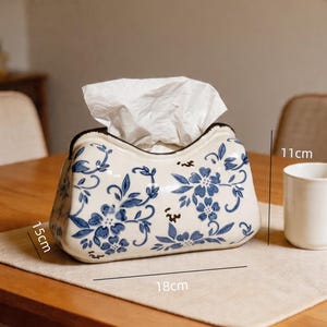 Op de afbeelding: Een keramische tissuebox met een blauw bloemmotief op een witte basis. De doos is 18 cm lang, 11 cm hoog en 15 cm breed. Witte tissues steken uit de bovenkant. Rechts staat een witte mok.