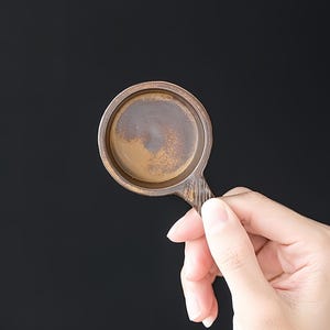 May include: A small, round, brown ceramic tea cup holder with a handle. The holder has a textured surface and a dark brown interior. The handle has vertical lines.
