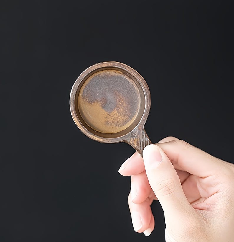 May include: A small, round, brown ceramic tea cup holder with a handle. The holder has a textured surface and a dark brown interior. The handle has vertical lines.