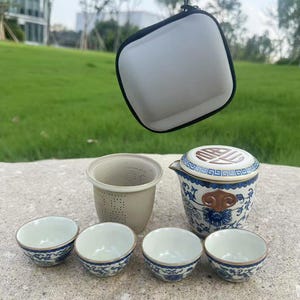 May include: A ceramic tea set with blue and white floral patterns, including a teapot, a strainer, and four small teacups. A white carrying case with a black trim is suspended above the set. The set is on a stone surface.