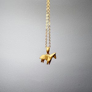 May include: A gold-colored necklace with a pendant shaped like a geometric horse. The pendant is suspended from a delicate chain. The background is a soft, neutral gray.