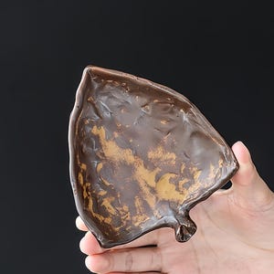 May include: A handcrafted, leaf-shaped dish with a textured, bronze-colored finish and gold accents. The small decorative bowl is held in a hand against a dark background. The dish is likely made of ceramic or metal.