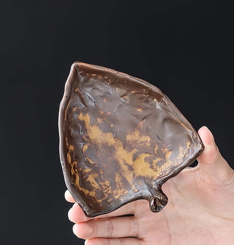 May include: A handcrafted, leaf-shaped dish with a textured, bronze-colored finish and gold accents. The small decorative bowl is held in a hand against a dark background. The dish is likely made of ceramic or metal.