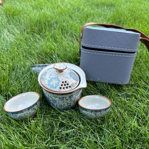 Pode incluir: Um conjunto de chá de cerâmica com um design floral em azul e dourado, incluindo uma chaleira, duas chávenas de chá e uma mala de transporte. A chaleira tem uma tampa e um coador embutido. As chávenas e a chaleira têm uma borda castanha. A mala de transporte cinzenta tem uma pega castanha.