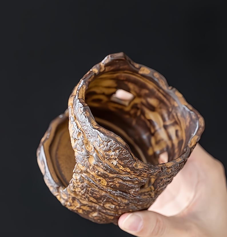 May include: A handcrafted, textured ceramic cup with a unique, organic design. The cup features a brown and tan color palette with a rough, bark-like exterior. The interior is smooth, contrasting the exterior's texture. The cup is held in a hand.