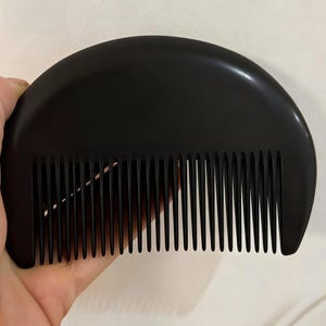 May include: A black, semi-circular comb with closely spaced teeth. The comb is held in a hand, showcasing its smooth, polished surface. The design is simple and functional, suitable for hair grooming.