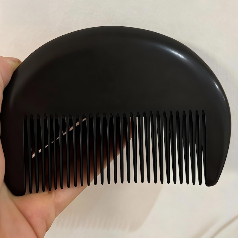 May include: A black, semi-circular comb with closely spaced teeth. The comb is held in a hand, showcasing its smooth, polished surface. The design is simple and functional, suitable for hair grooming.