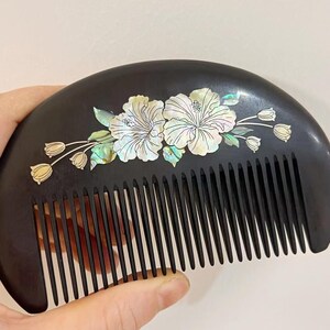 May include: A black wooden comb with a curved handle and fine teeth. The comb is decorated with two white hibiscus flowers and several small flower buds inlaid with iridescent mother-of-pearl.