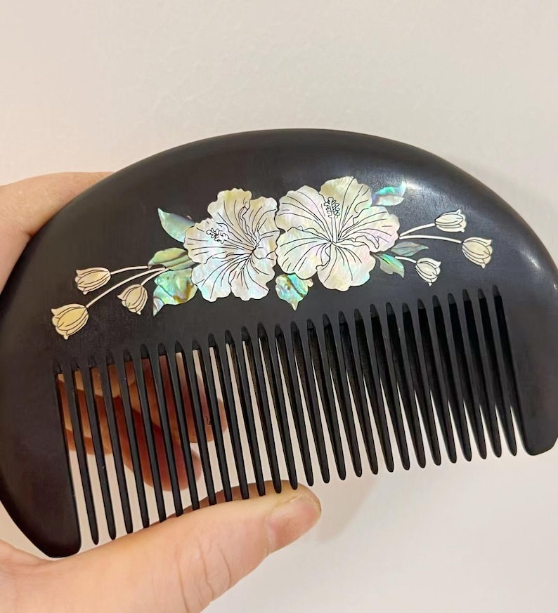 May include: A black wooden comb with a curved handle and fine teeth. The comb is decorated with two white hibiscus flowers and several small flower buds inlaid with iridescent mother-of-pearl.
