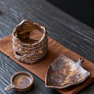 May include: A collection of handcrafted ceramic pieces in earthy tones. Includes a textured, cylindrical vase with cut-out details, a leaf-shaped dish, and a small cup with a handle. The items are arranged on a brown cloth.