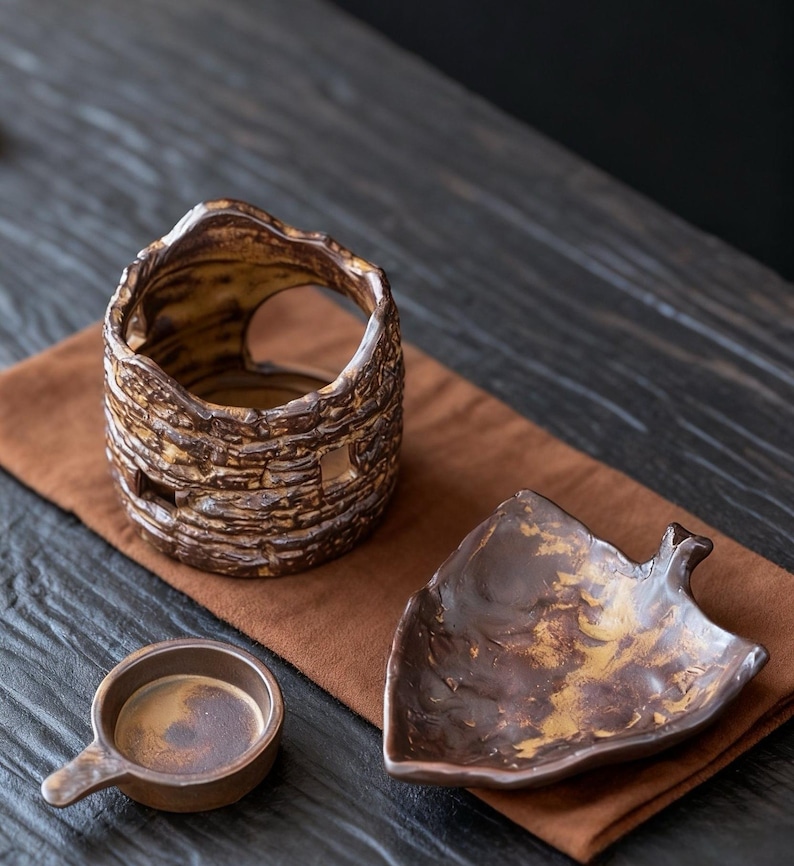 May include: A collection of handcrafted ceramic pieces in earthy tones. Includes a textured, cylindrical vase with cut-out details, a leaf-shaped dish, and a small cup with a handle. The items are arranged on a brown cloth.