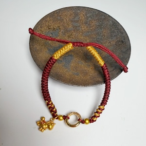 Può includere: Un braccialetto intrecciato rosso e oro con un ciondolo dorato e una chiusura ad anello dorato. Il braccialetto è realizzato in cordoncino intrecciato e presenta un piccolo ciondolo dorato. Il braccialetto è esposto su una superficie scura e rotonda.