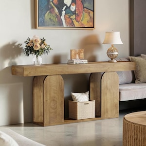 May include: A long, light brown wooden console table with two arched side supports and a lower shelf. The table holds a glass vase with beige flowers, a small wooden block, and two stacked books. A white woven basket sits on the lower shelf.