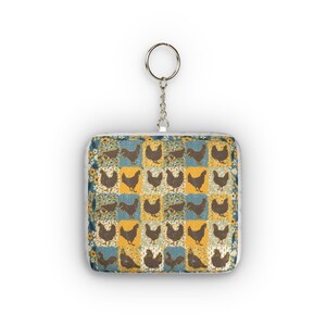 Può includere: Un portachiavi quadrato con un design patchwork. Presenta sagome di galline marroni su quadrati gialli e blu, con un bordo floreale. Il portachiavi ha un anello in metallo argentato.