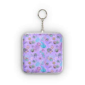 Könnte beinhalten: Ein quadratischer Schlüsselanhänger mit einem Meeresmuster. Das Design zeigt Schildkröten, Quallen, Seesterne und Muscheln in Rosa-, Blau- und Lilatönen auf einem lavendelfarbenen Hintergrund. Der Schlüsselanhänger hat einen silbernen Metallring.
