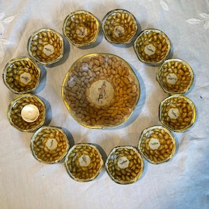 May include: A set of vintage peanut-themed serving dishes. The arrangement includes a large central bowl and several smaller, individual bowls, all featuring a peanut design. The bowls have a gold trim and are arranged on a white surface.