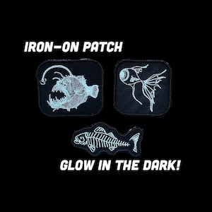 May include: Three black iron-on patches featuring white embroidered designs of an anglerfish, a fish eye, and a fish skeleton. The text IRON-ON PATCH and GLOW IN THE DARK! is printed in white. These patches have a square or contoured shape.