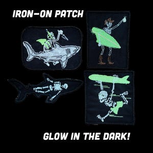 Könnte beinhalten: Vier schwarze Aufbügler mit gestickten Skelettmotiven. Eines zeigt ein Skelett auf einem Hai, eines mit einem Surfbrett, eines in Hai-Form und eines trägt ein Surfbrett. Der Text lautet: IRON-ON PATCH GLOW IN THE DARK!