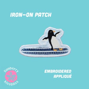 Op de afbeelding: Een opstrijkbare patch met een geborduurde pinguïn op een donkerblauw surfboard met witte strepen. De patch heeft een witte rand en de tekst IRON-ON PATCH en EMBROIDERED APPLIQUÉ en een roze bloemlogo zijn in de afbeelding zichtbaar.
