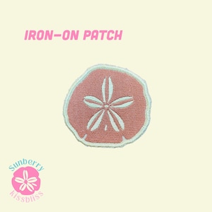 Puede incluir: Un parche termoadhesivo con un diseño circular de dólar de arena en rosa empolvado con detalles bordados en blanco. El texto IRON-ON PATCH aparece en rosa en la parte superior y un pequeño logotipo de dólar de arena rosa con Sunberry kissbliss abajo.