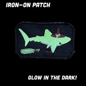 May include: A rectangular iron-on patch featuring an embroidered light green shark wearing a brown witch hat and riding a broomstick. The text IRON-ON PATCH and GLOW IN THE DARK! is printed in white against a solid black background.