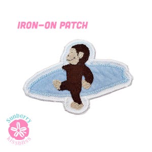 May include: An iron-on patch featuring a brown monkey character standing on a light blue surfboard. The patch has a white embroidered border and a white background. The text IRON-ON PATCH is printed in pink above the embroidered design.