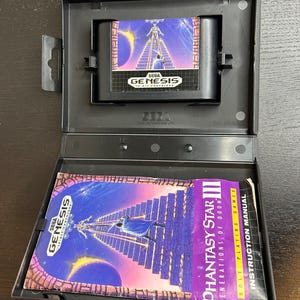 May include: A black Sega Genesis game cartridge case, open to reveal the game cartridge and instruction manual. The cartridge features a purple and blue graphic with the Sega Genesis logo. The instruction manual is titled "Phantasy Star III".