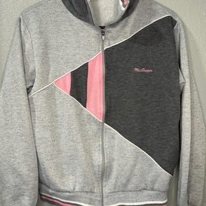 May include: A gray zip-up track jacket with a color block design. The jacket features a dark gray panel, a pink triangle, and white trim. The cuffs and hem have pink and white stripes. The brand name "MacGregor" is embroidered on the dark gray panel.