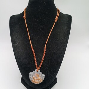 May include: A necklace featuring a semi-circular gray pendant with an embossed orange design, suspended from a thin, braided orange cord. The cord is knotted at the top and features a spiral macrame pattern leading down to the pendant.