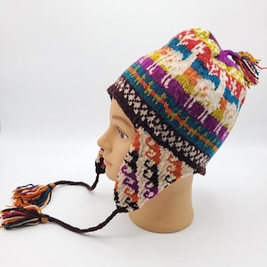 May include: A knit earflap hat featuring a colourful pattern of llamas and geometric shapes in shades of purple, blue, orange, and cream. The hat includes a braided cord with multi-coloured tassels on each side and a matching tassel on the crown.