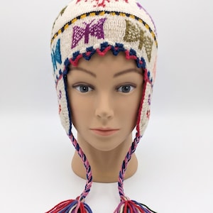May include: An off-white knit earflap hat featuring a colourful pattern of butterflies and birds. The hat has a scalloped dark blue and pink trim along the forehead and two long, braided ties ending in multi-coloured yarn tassels at the bottom.
