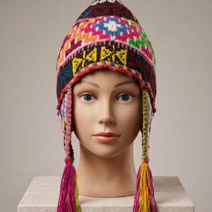 May include: A colorful knit hat featuring a pointed crown and ear flaps with braided ties ending in long tassels. The hat displays a complex geometric pattern in shades of maroon, bright pink, yellow, blue, green, and white across the entire surface.