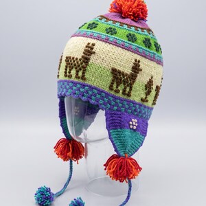 Puede incluir: Un gorro de punto con orejeras que presenta una banda de color crema con motivos de llamas marrones, acentuado por patrones geométricos en verde, morado y turquesa. El gorro tiene un gran pompón naranja y dos trenzas con pompones pequeños.