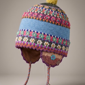 May include: A multi-coloured knit hat with earflaps and a yellow pom-pom on top. The hat features horizontal bands of floral patterns in pink, light blue, white, and dark grey, with braided tassels hanging from the sides and a scalloped edge.
